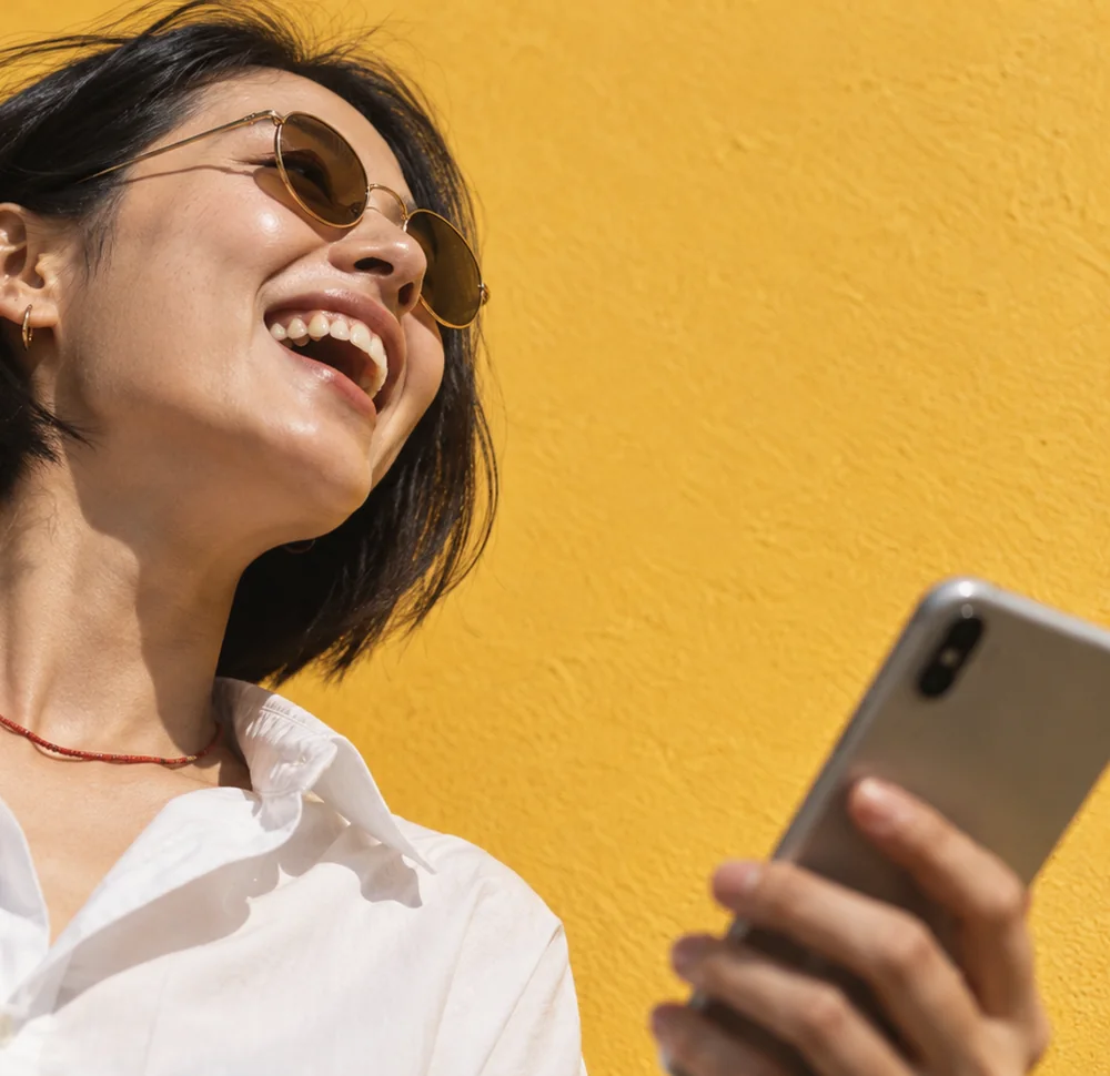 A woman in sunglasses laughs by a yellow wall, holding her phone
